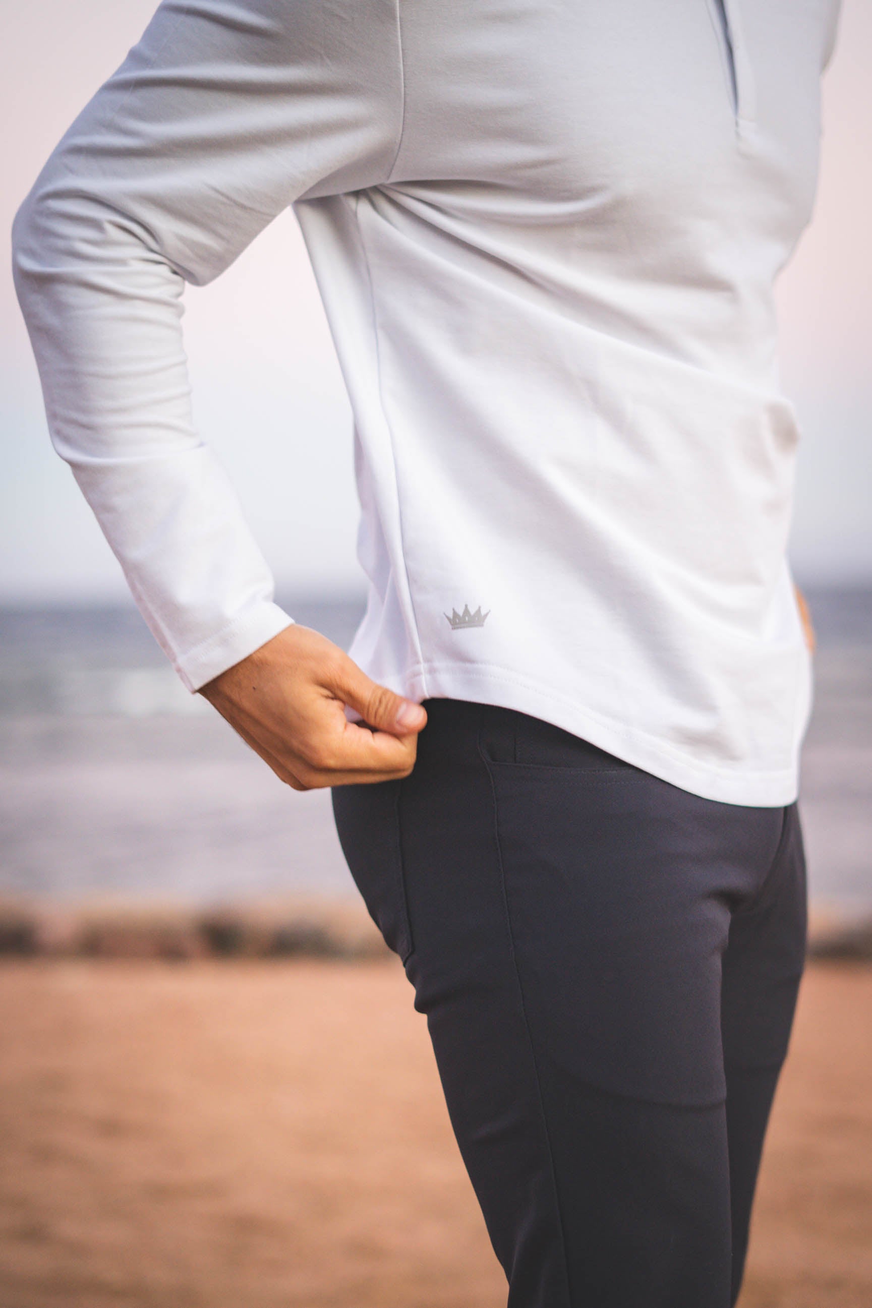 A person wearing a white long-sleeve flex shirt and dark pants, standing outdoors near a beach with a body of water in the background.