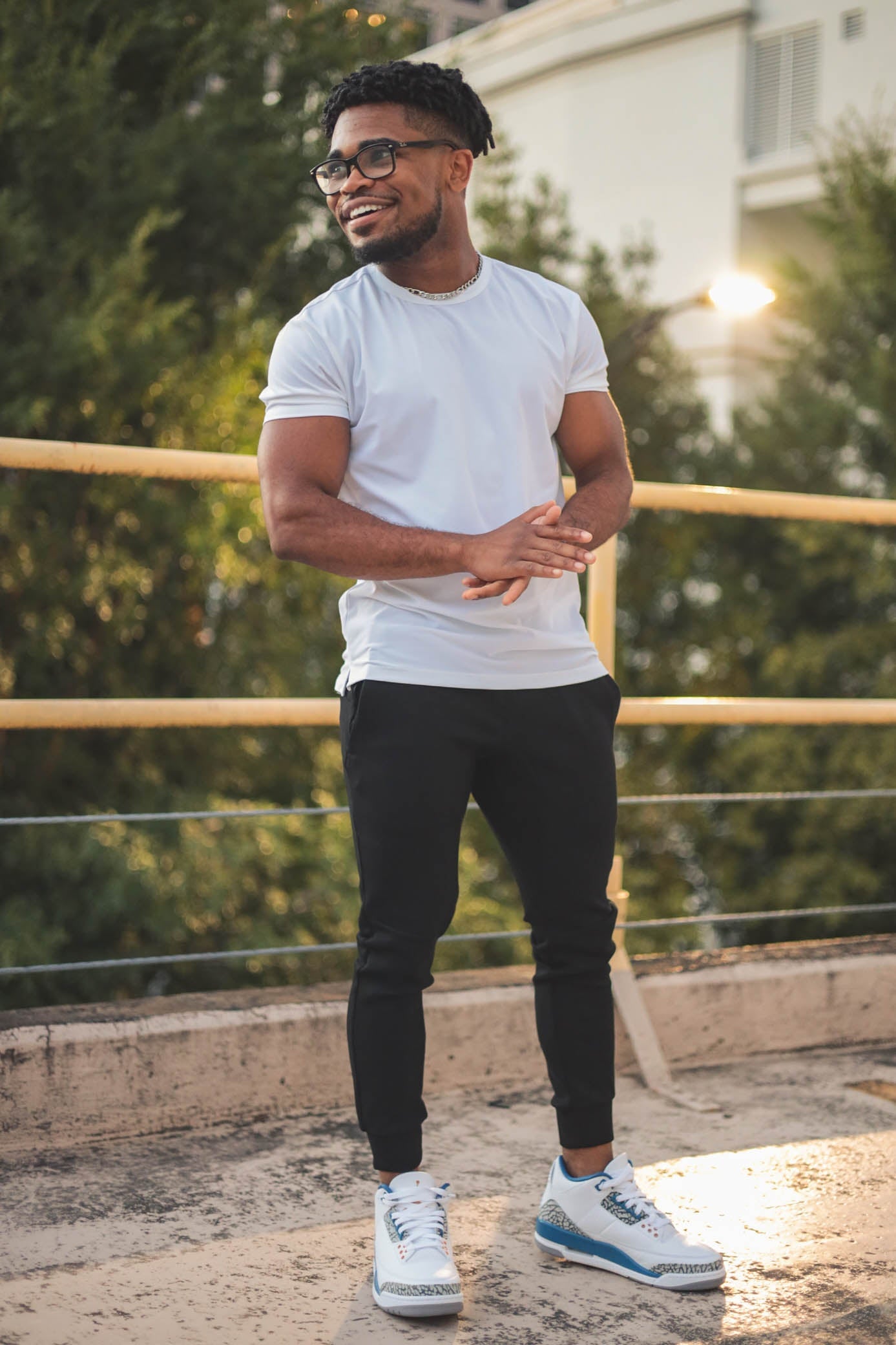 A man wearing glasses is smiling and standing outdoors. He is dressed in a white t-shirt, black jogger pants for short men from under510.com, and white sneakers with blue accents. Trees and a building are visible in the background.