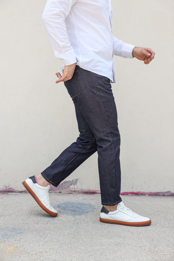 A person wearing dark denim xavier jeans from under510.com, a white button-up shirt, and white sneakers with brown soles is walking on a concrete surface.