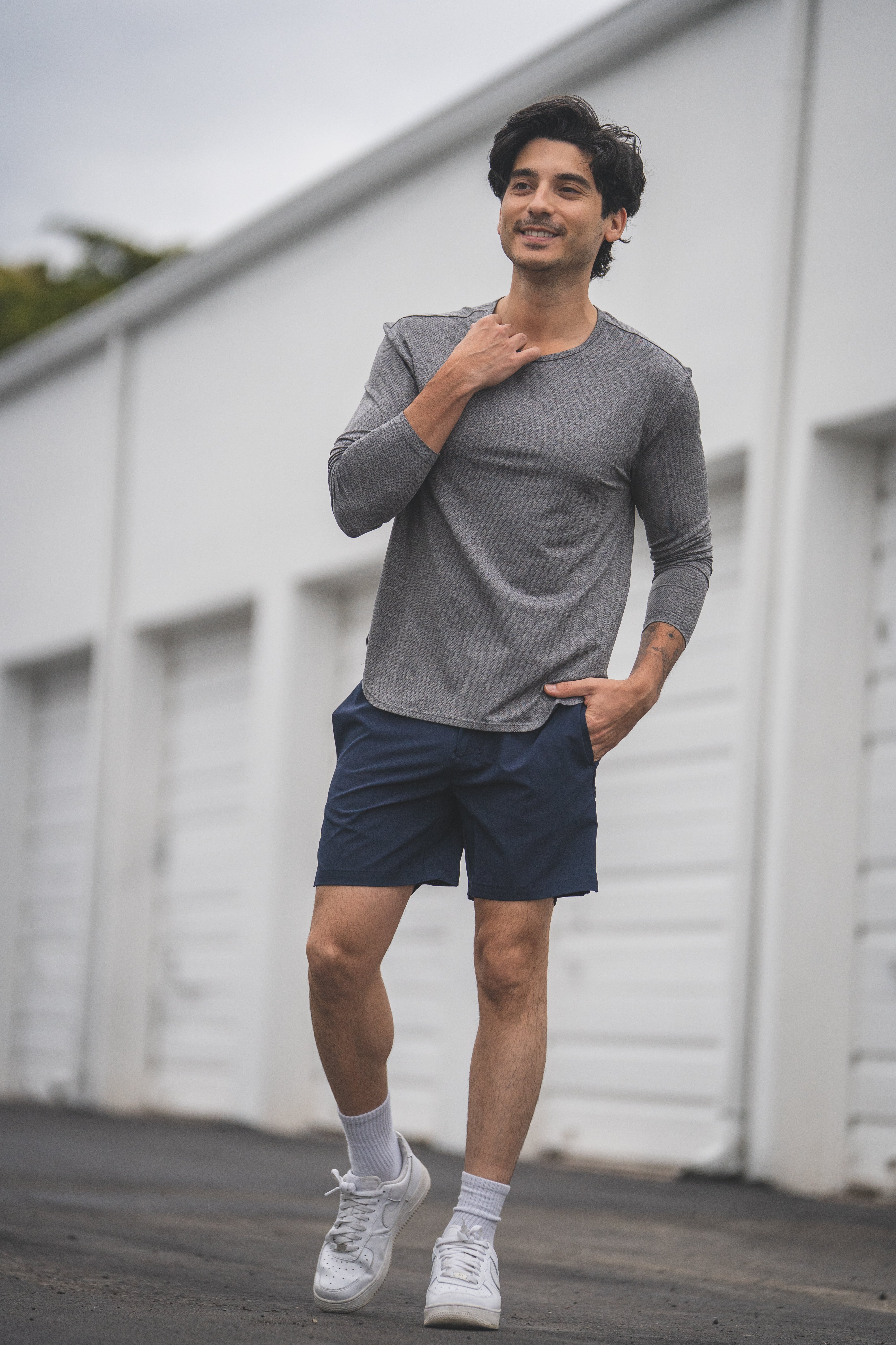 Man wearing a gray long-sleeve shirt and navy shorts standing in front of a white building.