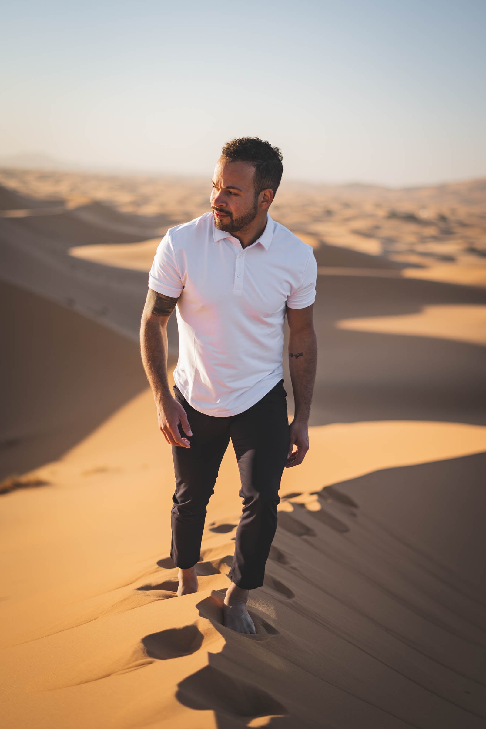 Male model walking on sand dune in egypt featuing under510.com clothing such as a white flex polo and black super stretch performance pants
