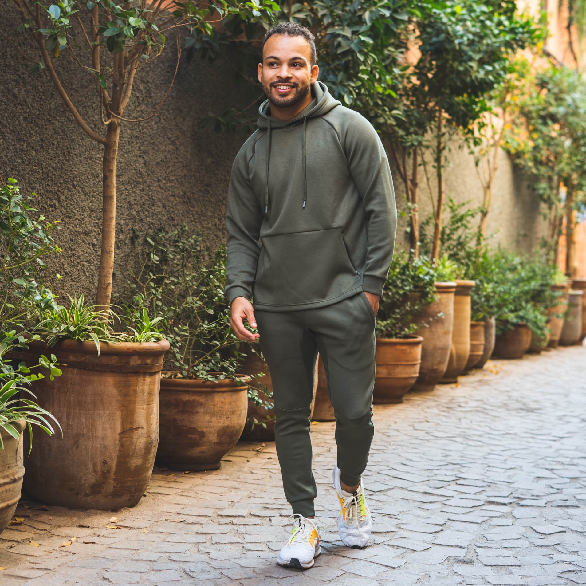 The image shows a man walking outdoors in a narrow pathway lined with potted plants. He is wearing an olive green hoodie and matching jogger pants, paired with white sneakers featuring yellow accents. The man has a cheerful expression and is walking with his hands in his pockets. The background includes a wall with greenery and tall plants in large terracotta pots, creating a serene and natural atmosphere.