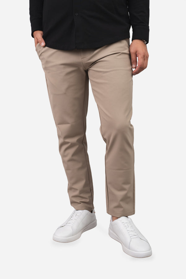 A person wearing slim-fit beige chinos with a black long-sleeve shirt and white sneakers, shown from the waist down against a light gray studio background.