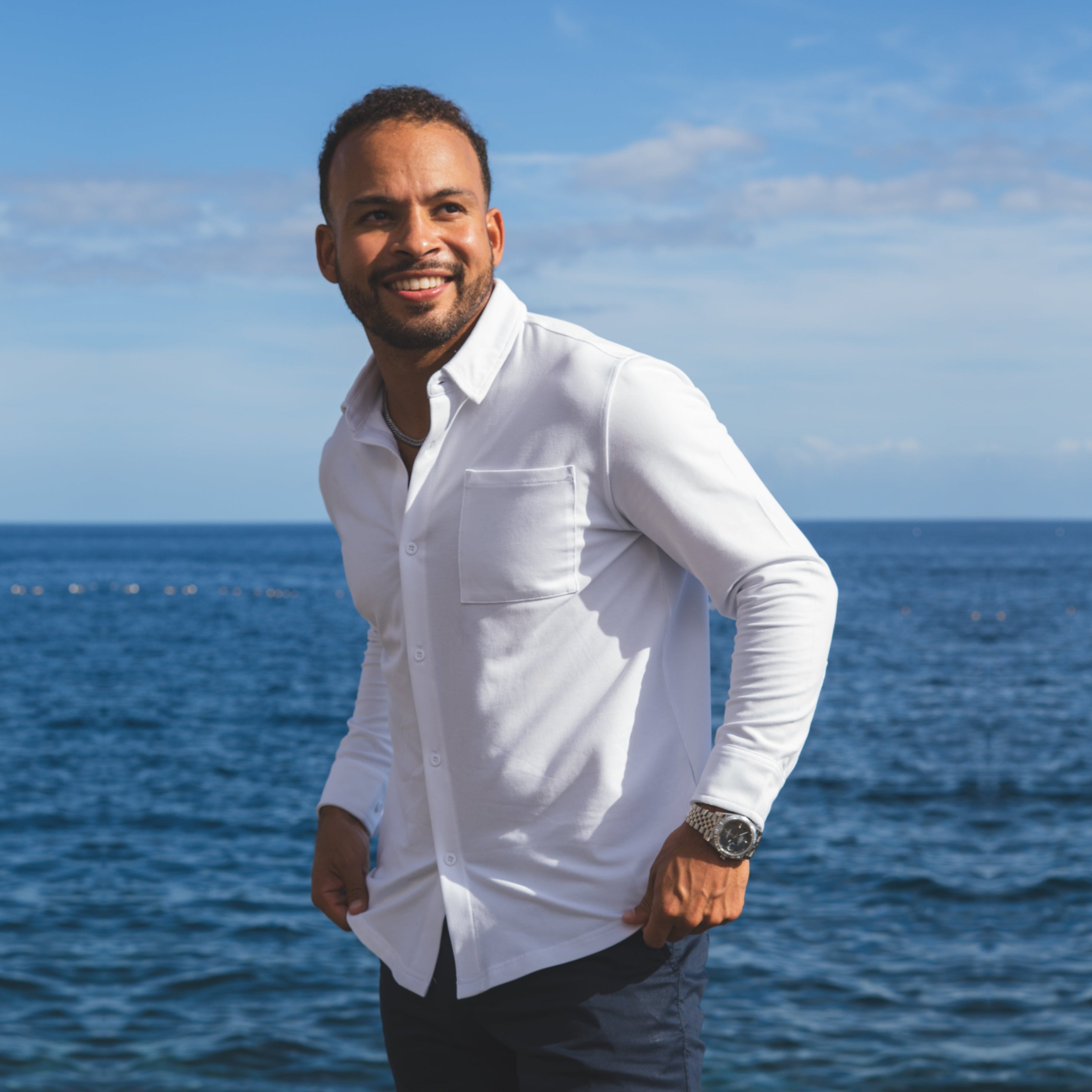 Man in a white shirt standing by the ocean with a clear blue sky.
